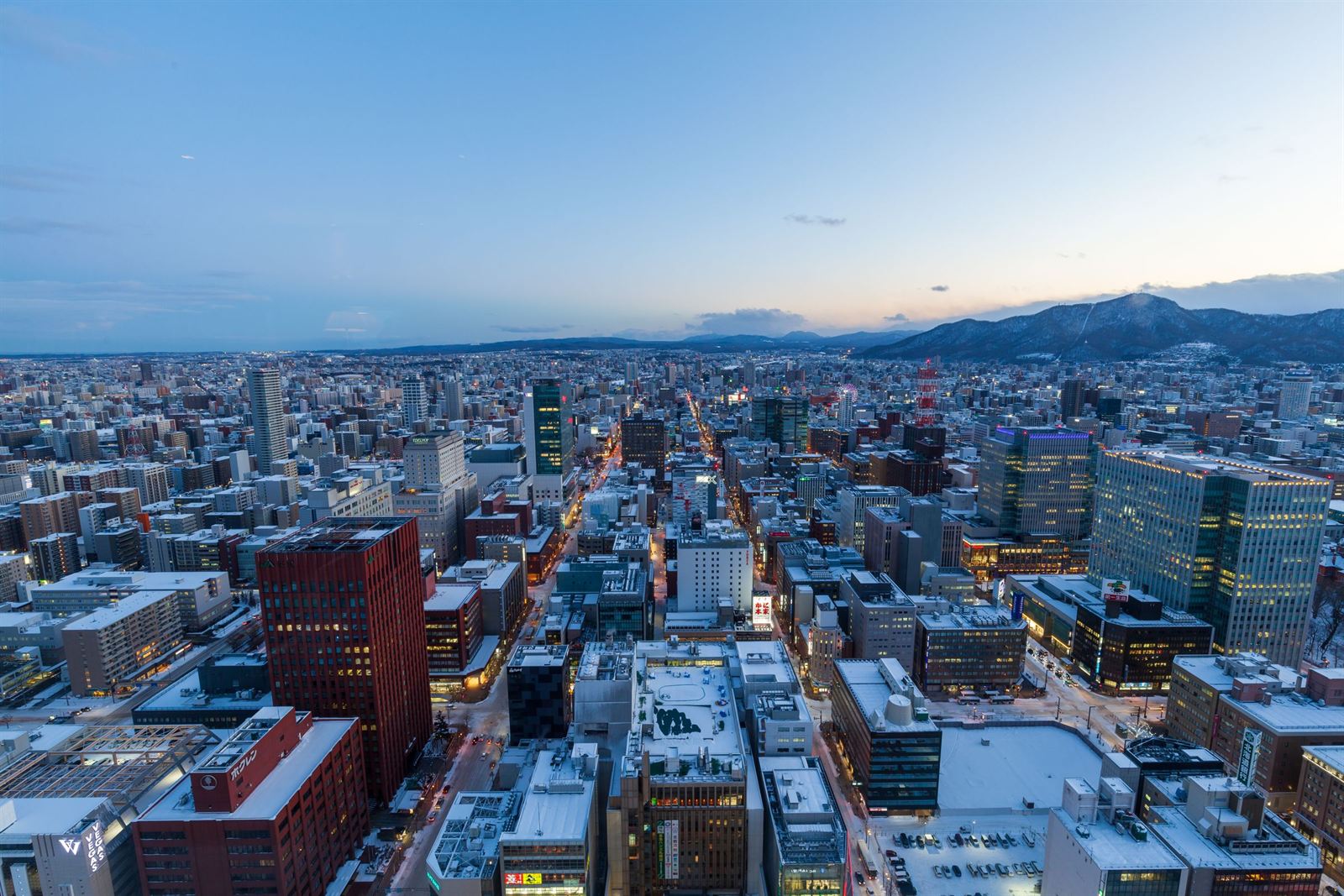 Elevated winter view over snowy Sapporo's grid of city blocks toward mountains at dusk