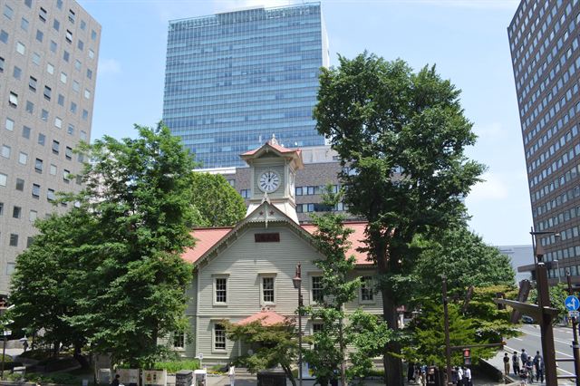 Historic white wooden Sapporo Clock Tower among trees and modern office towers