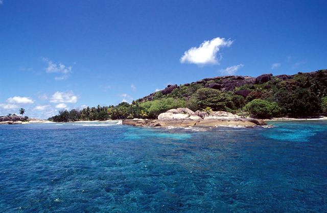 Granite outcrops and lush green island shore above clear blue water in La Digue, Seychelles