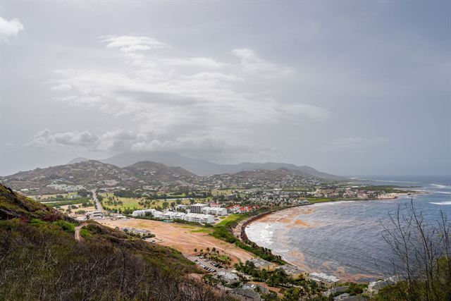 Elevated view over St Kitts coastline with a beach, low hills and a resort area under cloudy sky