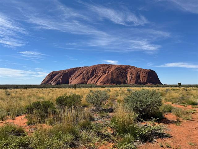 Uluru's red monolith rising above desert scrub and spinifex under a streaked blue sky