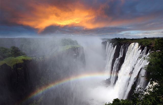 Victoria Falls plunging into its gorge with a rainbow in the spray under a sunset sky